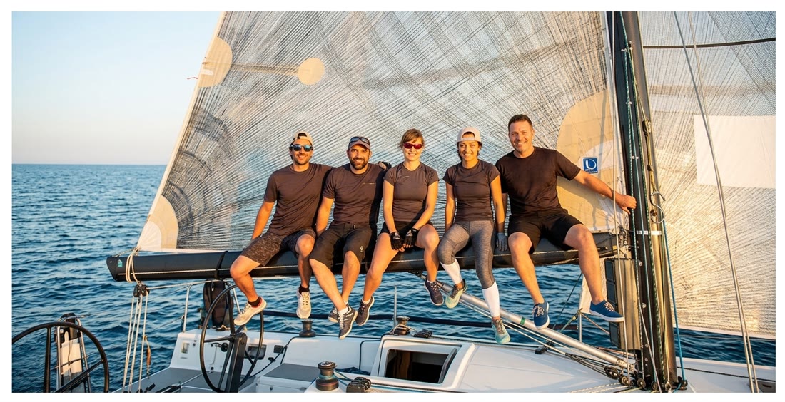 Five people sitting on the boom of a sailing boat in the evening sun